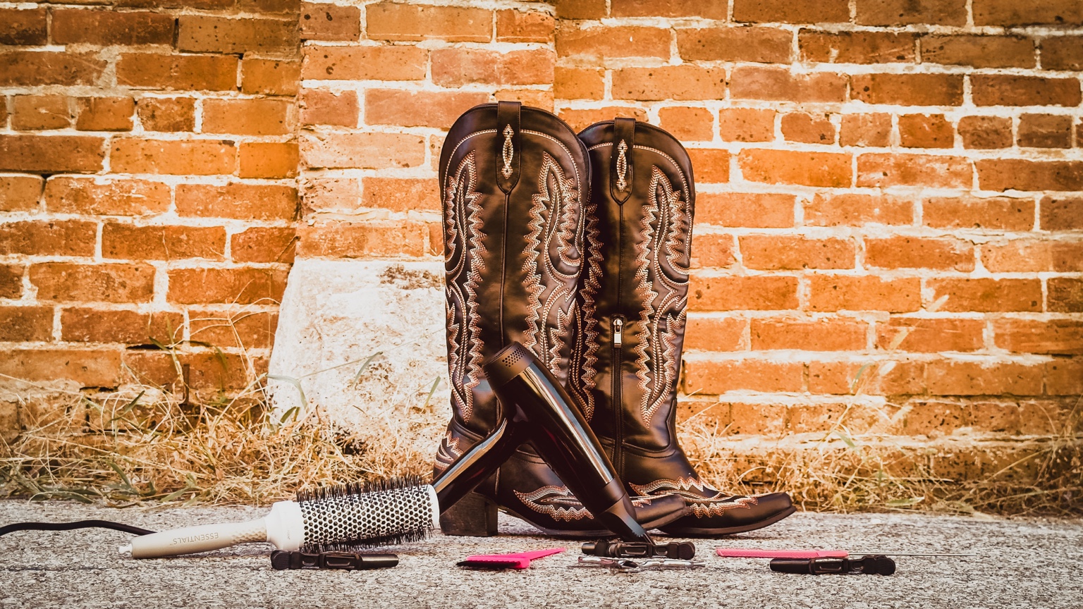 Embroidered western boots, a round brush, and pink hair tools styled against warm brickwork